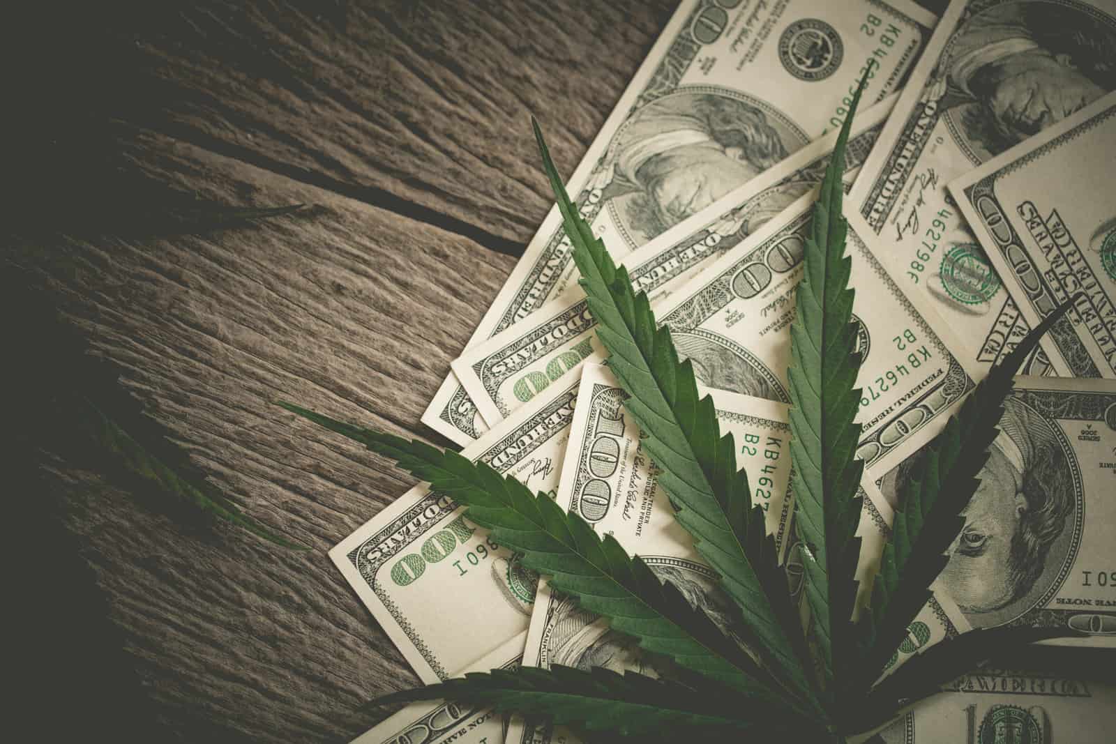 Coronavirus Federal Stimulus Bills: FAQ For Marijuana Businesses. $100 bills and marijuana leaves on wood surface.